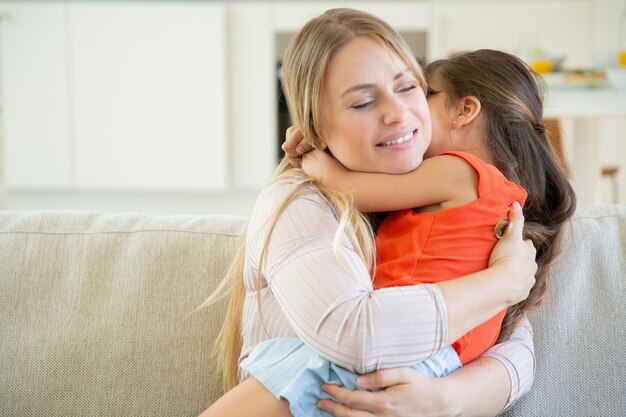 Friedliche Mutter hält ihr kleines Mädchen in den Armen auf ihrem Schoß und umarmt sie.