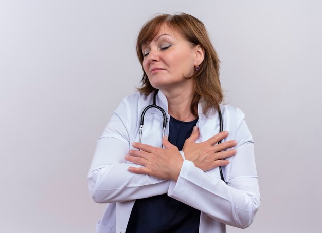 Friedliche Ärztin mittleren Alters, die medizinische Robe und Stethoskop trägt, die Hände auf Brust mit geschlossenen Augen auf isolierter weißer Wand mit Kopienraum setzen