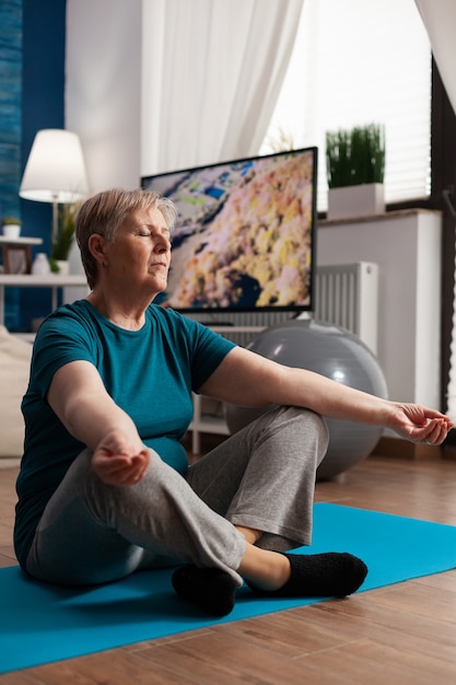 Friedliche Seniorin sitzt bequem im Lotussitz auf Yogamatte mit geschlossenen Augen