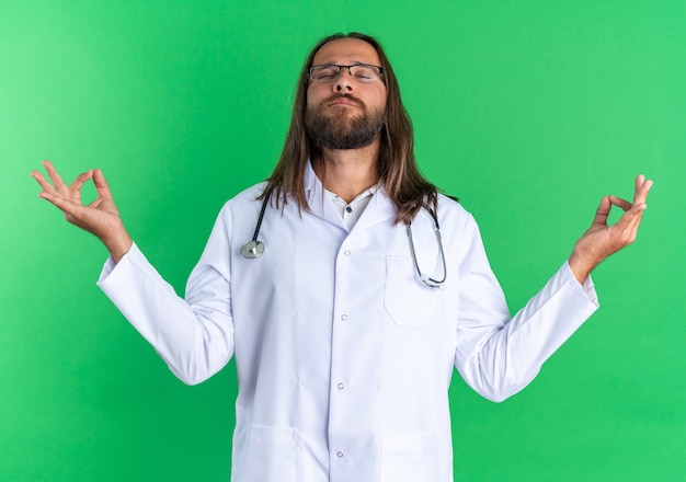 Friedlicher erwachsener männlicher Arzt mit medizinischem Gewand und Stethoskop mit Brille, der mit geschlossenen Augen meditiert, isoliert auf grüner Wand