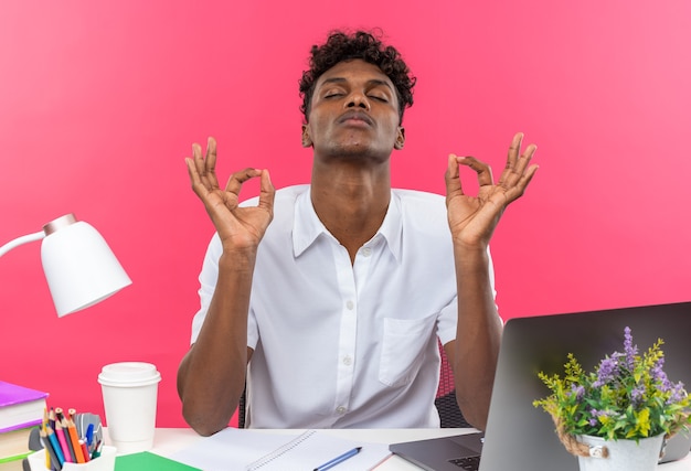 Friedlicher junger afroamerikanischer Student, der am Schreibtisch mit Schulwerkzeugen sitzt und isoliert auf rosa Wand meditiert
