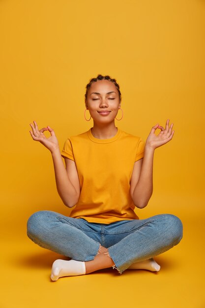 Friedliches, ruhiges, dunkelhäutiges weibliches Model sitzt mit gekreuzten Beinen auf dem Boden, praktiziert Yoga und versucht sich zu entspannen, atmet tief durch, schließt die Augen, erreicht das Nirvana, hält die Hände seitwärts und löst nach der Arbeit Stress ab