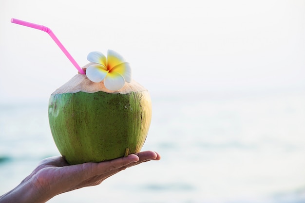 Frische Kokosnuss in der Hand mit dem Plumeria verziert auf Strand mit Seewelle - Tourist mit Sonnenferienkonzept der frischen Frucht und des Meersands