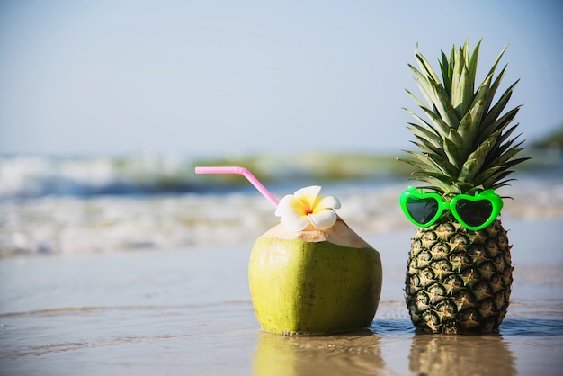 Frische Kokosnuss und Ananas setzten reizende Gläser der Sonne auf sauberen Sandstrand mit Seewelle - frische Frucht mit Meersandsonnen-Ferienkonzept