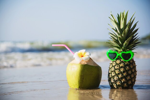 Frische Kokosnuss und Ananas setzten reizende Gläser der Sonne auf sauberen Sandstrand mit Seewelle - frische Frucht mit Meersandsonnen-Ferienkonzept