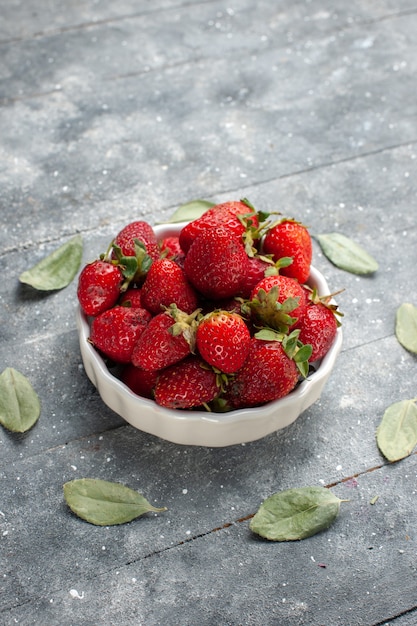 Kostenloses Foto frische rote erdbeeren in weißer platte zusammen mit grünen getrockneten blättern auf grauem schreibtisch