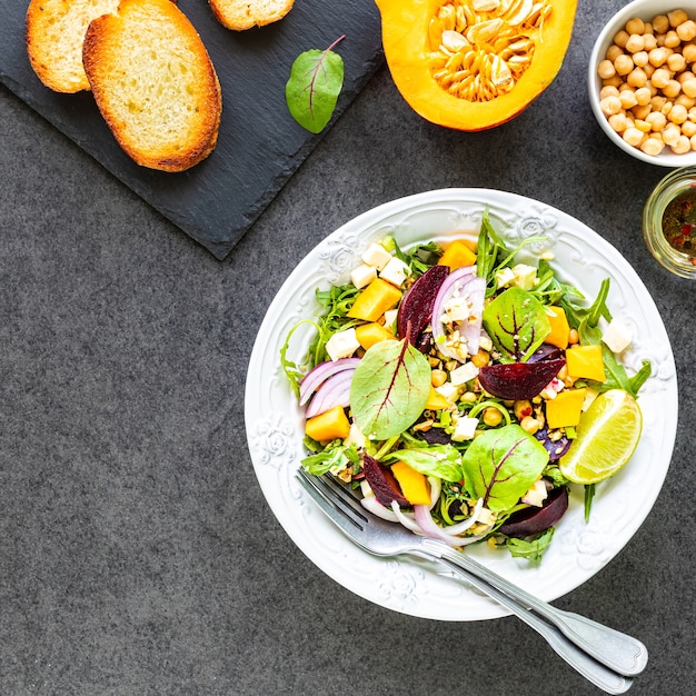 Frischer Gemüsesalat mit Rüben, Rucola, roten Zwiebeln und Sauerampfer in einem weißen Teller mit Kürbis, Toast und Kichererbsen