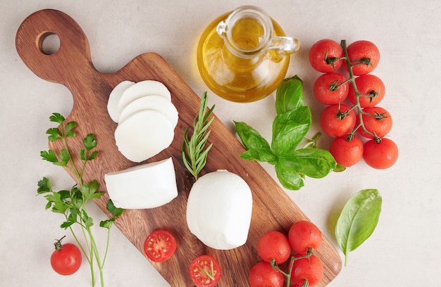 Frischer Mozzarella-Käse, italienischer Weichkäse, Tomaten und Basilikum, Olivenöl und Rosmarin auf hölzernem Servierbrett über heller Holzoberfläche. Gesundes Essen. Draufsicht. Flach liegen.