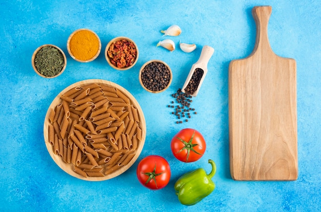 Frisches Bio-Gemüse mit roher Pasta und Gewürzen. Holzbrett auf blauem Hintergrund.