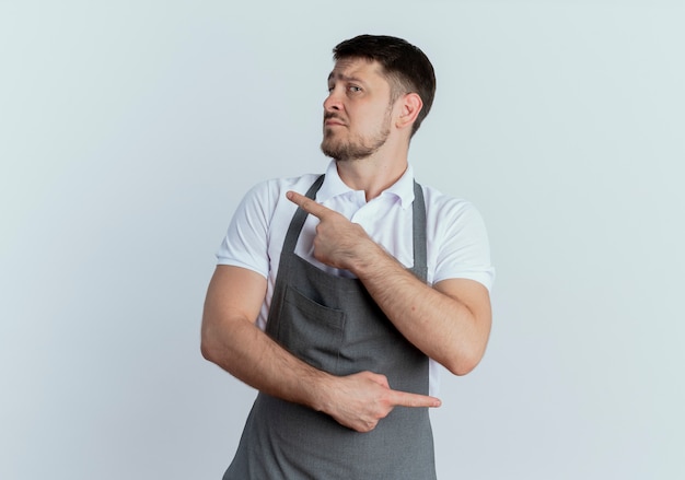 Friseur in der Schürze, die mit ernstem Gesicht beiseite schaut, das mit Zeigefingern zu verschiedenen Richtungen zeigt, die über weißer Wand stehen