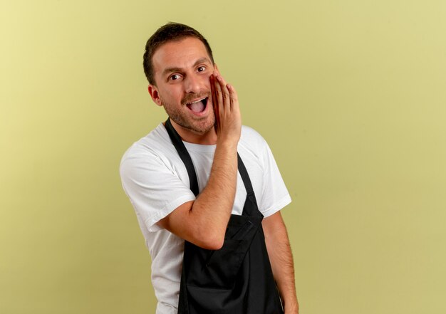 Friseur in der Schürze, die nach vorne schaut und ein Geheimnis mit der Hand nahe dem Mund erzählt, der über der hellen Wand steht