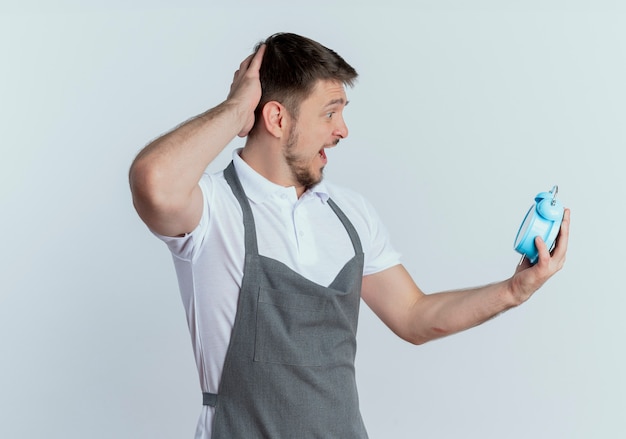 Friseur in der Schürze hält Wecker, der sie mit der Hand auf seinem Kopf verwirrt ansieht, vergessen, schlechtes Gedächtniskonzept, das über weißer Wand steht