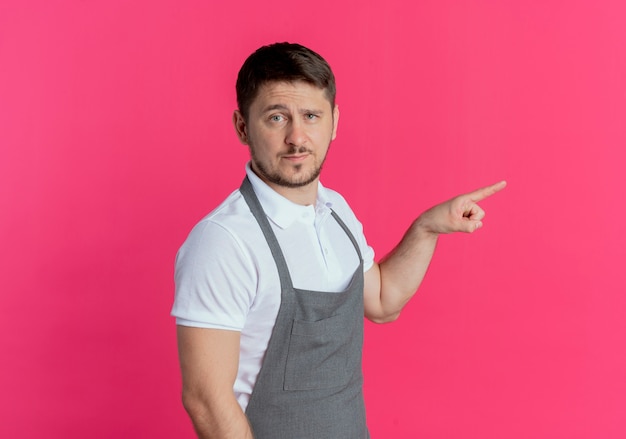 Friseur in der Schürze mit dem selbstbewussten Ausdruck, der mit dem Finger zur Seite zeigt, die über rosa Wand steht