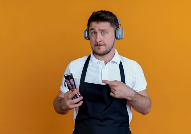 Friseur in der Schürze mit Kopfhörern, die Bartschneider und Schere halten, die mit dem Finger auf sie zeigen, lächelnd stehend über orange Wand