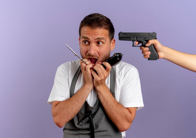 Friseur in Schürze mit Schere und Trimmer erschrocken, während ihn jemand mit einer Waffe anvisiert