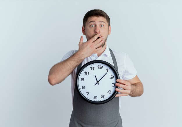 Friseur Mann in der Schürze hält Wanduhr, die Mund mit Hand bedeckt, die über weißer Wand geschockt steht