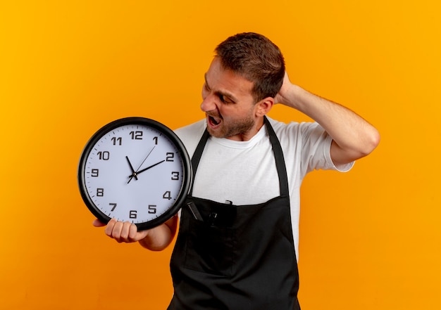 Friseur Mann in der Schürze hält Wanduhr und schaut sie verwirrt und sehr besorgt über orange Wand stehen