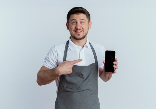 Friseur Mann in der Schürze zeigt Smartphone mit dem Finger darauf lächelnd über weiße Wand stehend