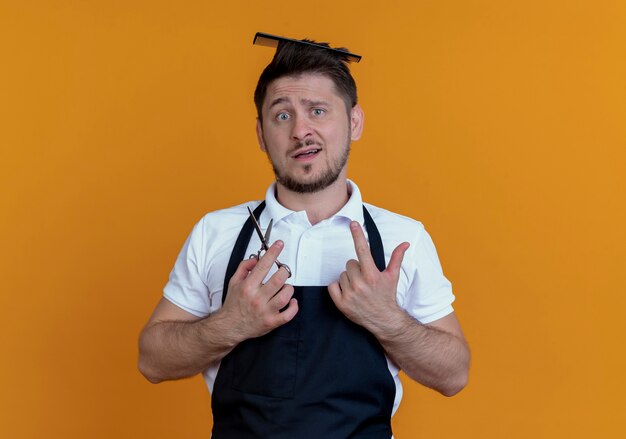 Kostenloses Foto friseur mann in schürze mit haarbürste in seinem haar hält schere verwirrt über orange wand stehen