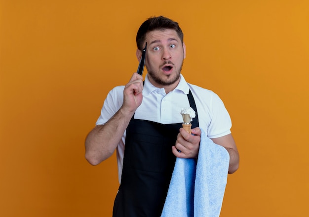 Friseur Mann in Schürze mit Handtuch auf der Hand hält Rasierpinsel mit Schaum und Rasiermesser verwirrt vergessen etwas Wichtiges über der orangefarbenen Wand stehen