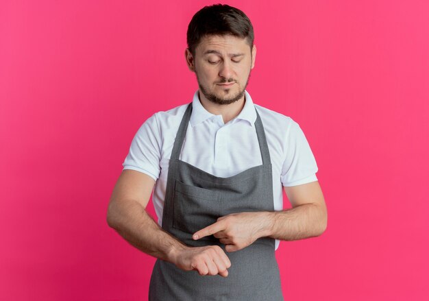 Friseurmann in der Schürze, der auf seinen Arm zeigt, erinnert an Zeit mit ernstem Gesicht, das über rosa Hintergrund steht