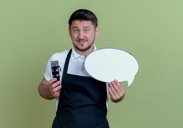 Friseurmann in der Schürze, die Bartschneider und leeres Sprachblasenzeichen hält, das mit glücklichem Gesicht über grüner Wand steht