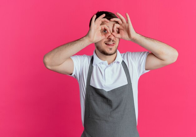 Friseurmann in der Schürze, die binokulare Geste mit den Fingern macht, die durch lächelnde Finger schauen, die über rosa Wand stehen