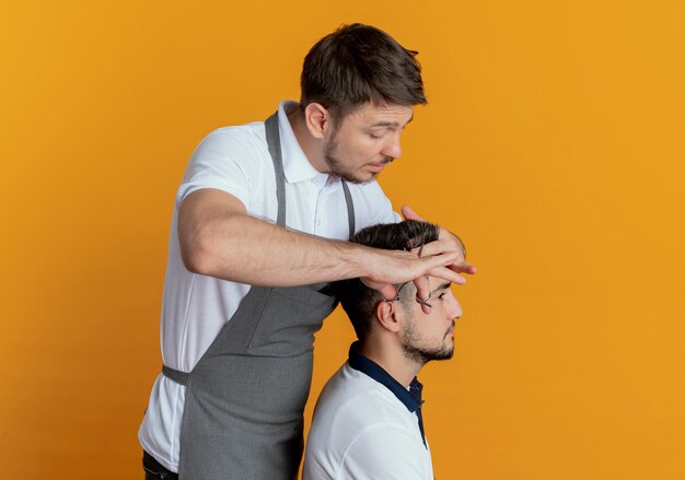 Friseurmann in der Schürze, die Haar mit Schere des zufriedenen Kunden über orange Hintergrund schneidet