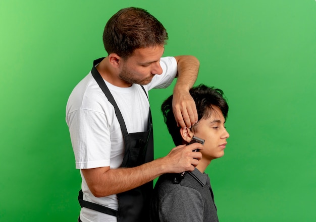 Friseurmann in der Schürze, die Haare mit der Schere des zufriedenen Kunden schneidet, der über grüner Wand 2 steht