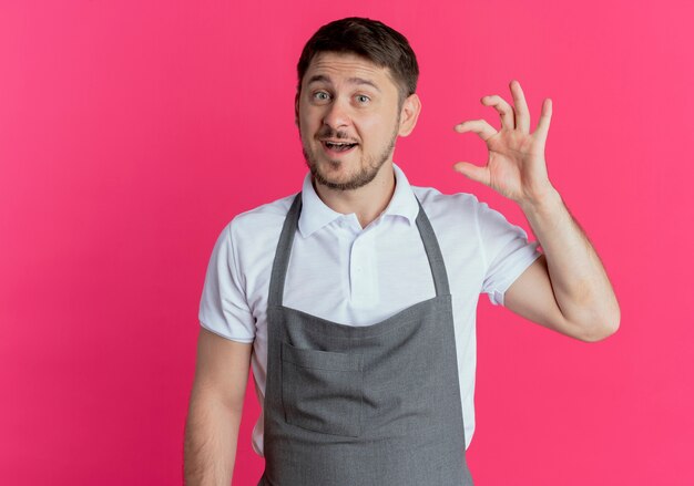 Friseurmann in der Schürze, die kleines Zeichen mit den Fingern zeigt, Maßsymbol, das über rosa Wand steht