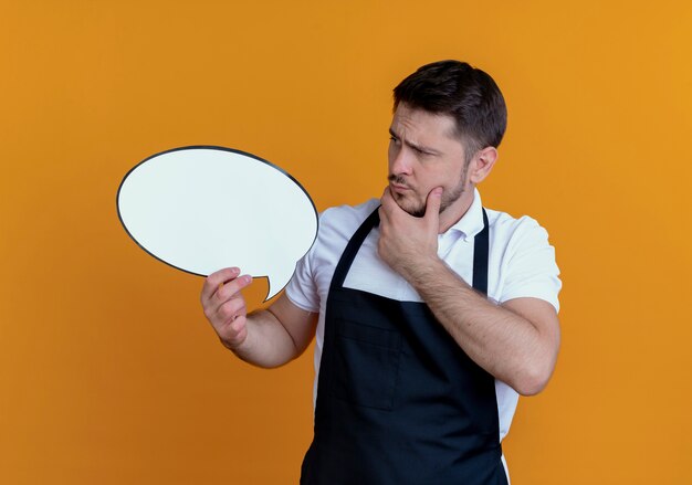 Friseurmann in der Schürze, die leeres Sprachblasenzeichen hält, das es betrachtet, das über orange Hintergrund steht