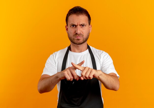 Friseurmann in der Schürze, die nach vorne mit stirnrunzelndem Gesicht schaut und Verteidigungsgeste kreuzt, die Finger über orange Wand steht