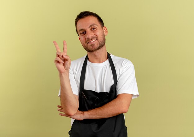 Friseurmann in der Schürze, die nach vorne schauend das Siegeszeichen zeigt, das fröhlich über der hellen Wand stehend lächelt