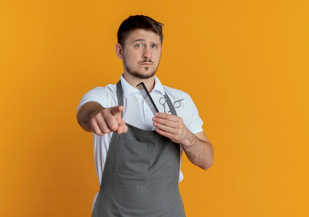 Friseurmann in der Schürze, die Schere hält, die mit dem Finger zur Kamera mit ernstem Gesicht zeigt, das über orange Hintergrund steht