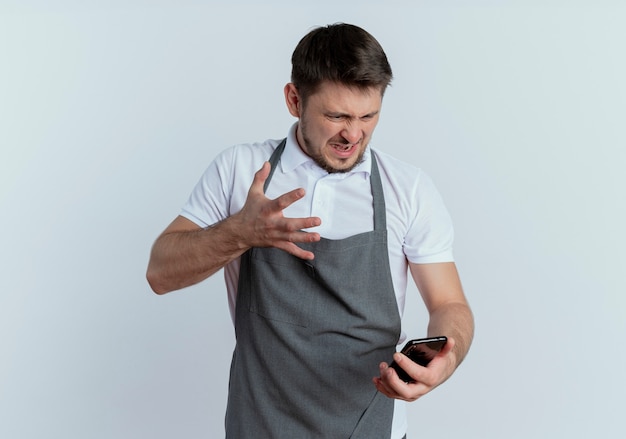 Friseurmann in der Schürze, die Smartphone hält, das Bildschirm mit genervtem Ausdruck betrachtet, der über weißem Hintergrund unzufrieden steht