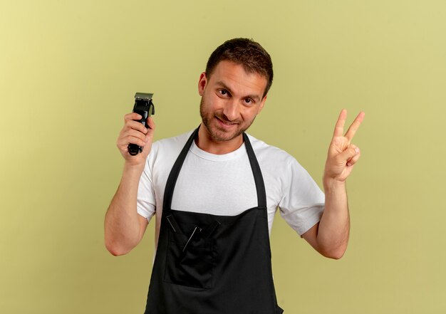 Friseurmann in der Schürze, die Trimmer hält, der Siegeszeichen zeigt, das fröhlich über heller Wand stehend lächelt