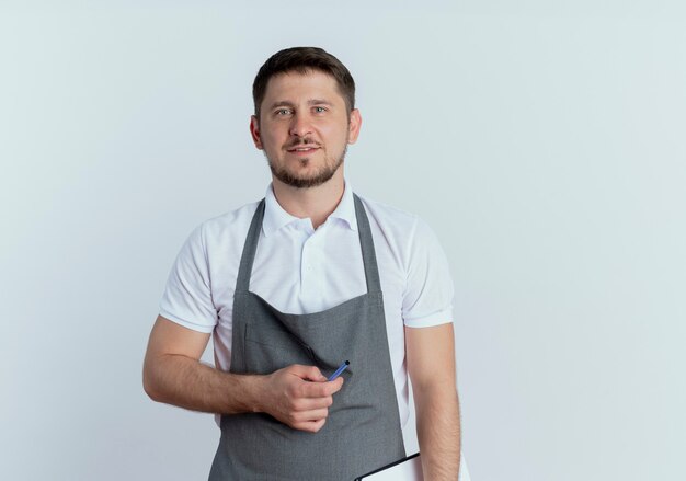 Friseurmann in der Schürze, die Zwischenablage und Stift mit Lächeln auf Gesicht hält, das über weißer Wand steht