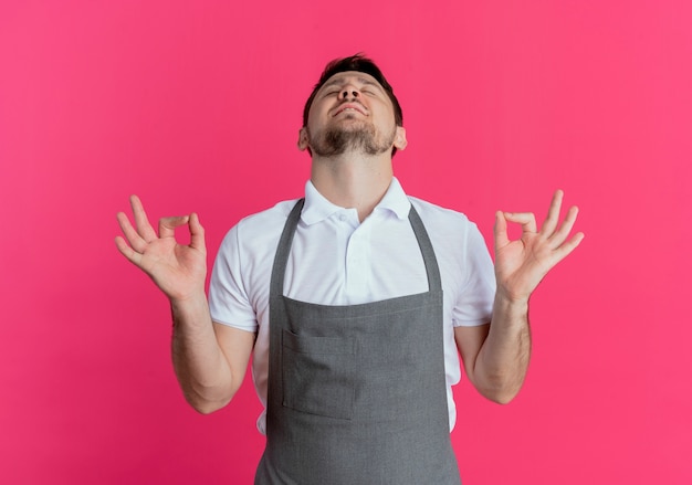 Kostenloses Foto friseurmann in der schürze entspannend, die meditationsgeste mit den fingern mit geschlossenen augen macht, die über rosa hintergrund stehen