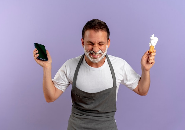 Friseurmann in der Schürze mit Rasierschaum auf seinem Gesicht, das Smartphone und Rasierpinsel hält, der aufgeregt und glücklich steht, über lila Wand stehend