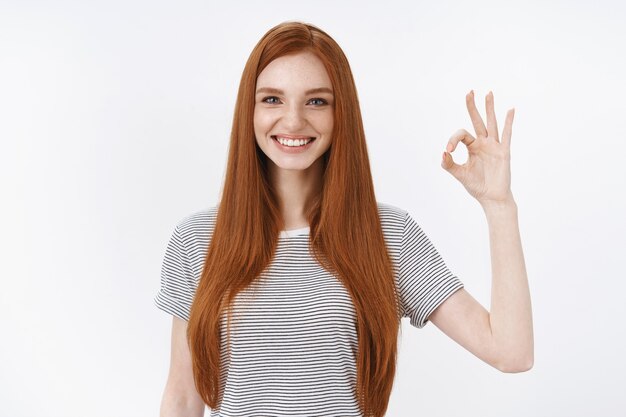 Fröhlich begeisterte attraktive rothaarige Studentin gibt positive Antwort zeigen okay ok normale Geste drückt positive gute Einstellung aus wie Ergebnis, stehende weiße Wand