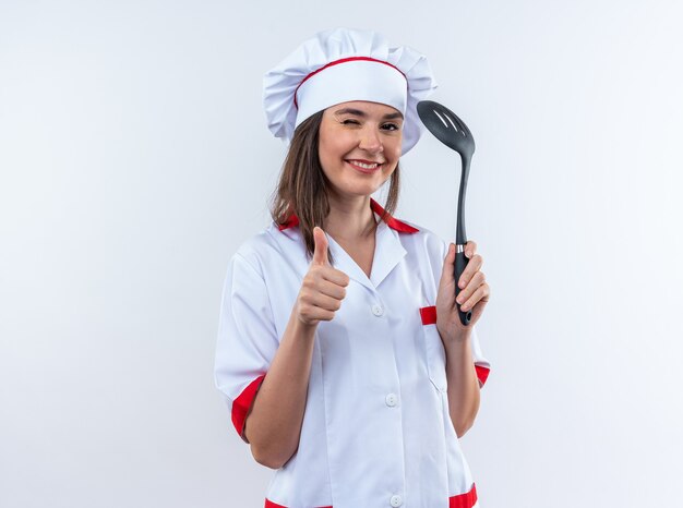 Fröhlich blinzelte junge Köchin in Kochuniform mit Spachtel, der Daumen nach oben isoliert auf weißem Hintergrund zeigt showing