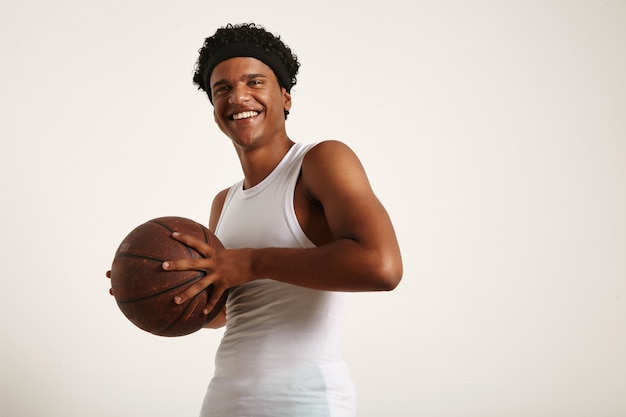 fröhlich lachender junger Afroamerikaner, der ein weißes ärmelloses Hemd und ein Stirnband webt, das einen Grunge-Lederbasketball an seine auf Weiß isolierte Brust hält.