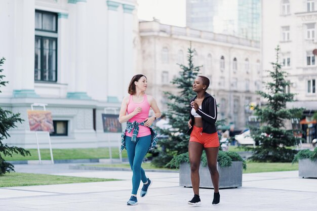 Fröhlich lächelnde Freunde in Sportbekleidung, die in der Stadt laufen und multiethnische Frauen beim Fitnesstraining diskutieren