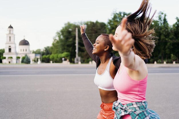 Fröhlich lächelnde Freunde in Sportbekleidung, die in der Stadt laufen und multiethnische Frauen beim Fitnesstraining diskutieren