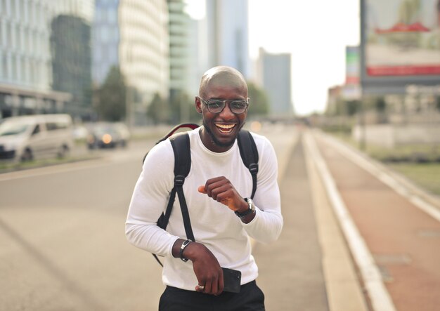 Fröhlich lächelnder afrikanischer Mann mit Brille, der ein weißes T-Shirt und einen Rucksack auf der Straße trägt