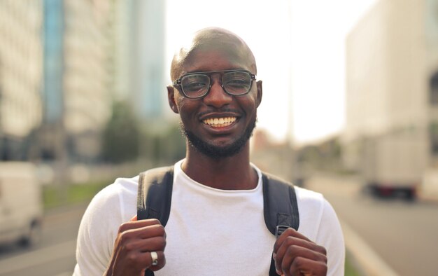 Fröhlich lächelnder afrikanischer Mann mit Brille, der ein weißes T-Shirt und einen Rucksack auf der Straße trägt