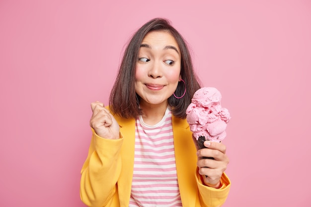 Fröhlich überraschte junge Asiatin sieht sich große appetitliche Eiscreme an und lächelt angenehm, genießt es, etwas leckeres Pausendiät zu essen, das in modischer Kleidung isoliert über rosa Wand gekleidet ist