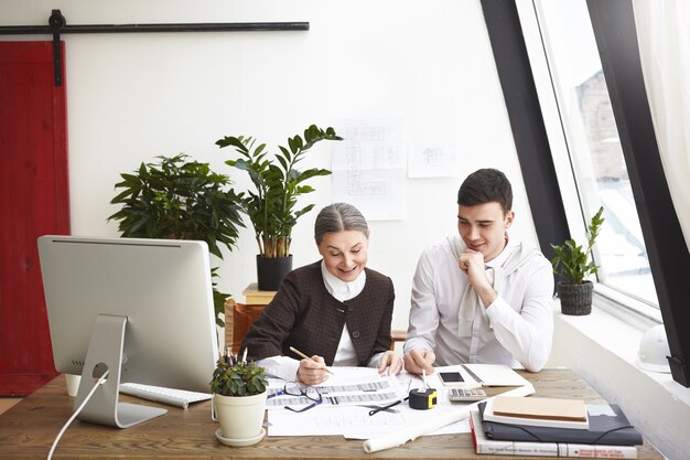 Fröhliche ältere Chefarchitektin und junger Mann, die gemeinsam an einem Architekturprojekt arbeiten, glücklich über die großartigen Ergebnisse ihrer gemeinsamen Arbeit, reden und lächeln. Teamwork und Zusammenarbeit