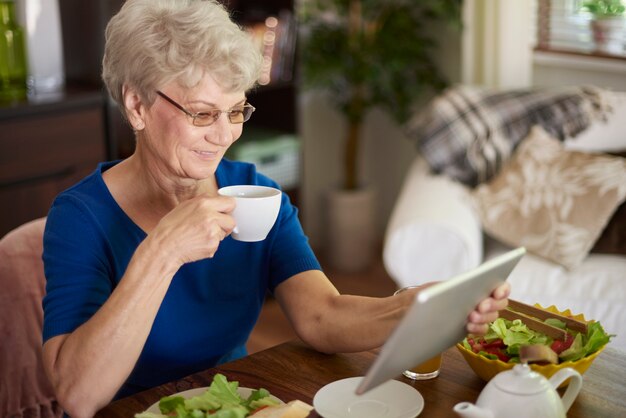 Fröhliche ältere Frau, die Frühstück isst und im Internet surft