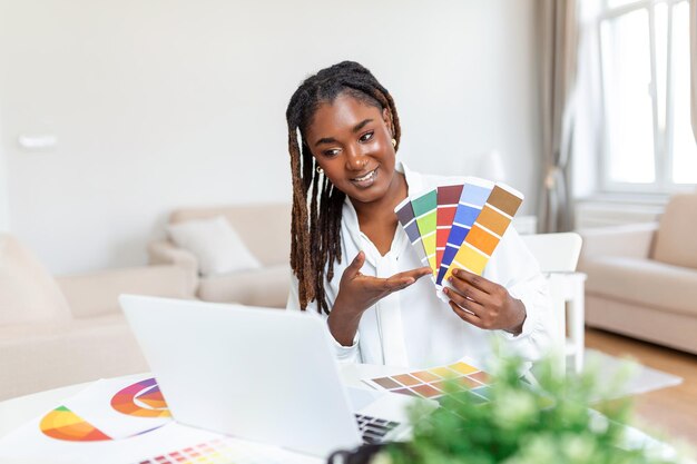 Fröhliche afrikanisch-amerikanische Designerin bei einer Videokonferenz mit Kunden, die am Schreibtisch vor dem Computer sitzen und Farbpaletten halten, gestikulierend und lächelnd Kopienraum haben